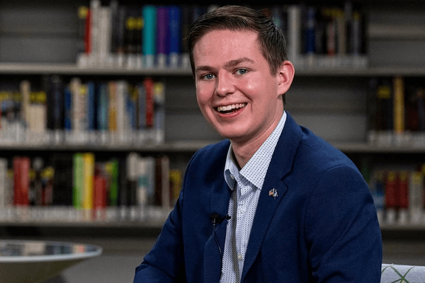 A smiling young man in a navy blue blazer and patterned white shirt sits in front of library bookshelves.  A small lavalier microphone is clipped to his lapel.